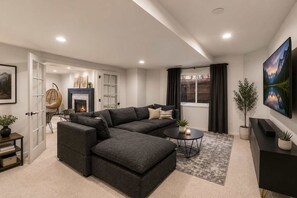 Un salon moderne avec un canapé d’angle, une cheminée, un téléviseur et une grande fenêtre agrémentée de rideaux.