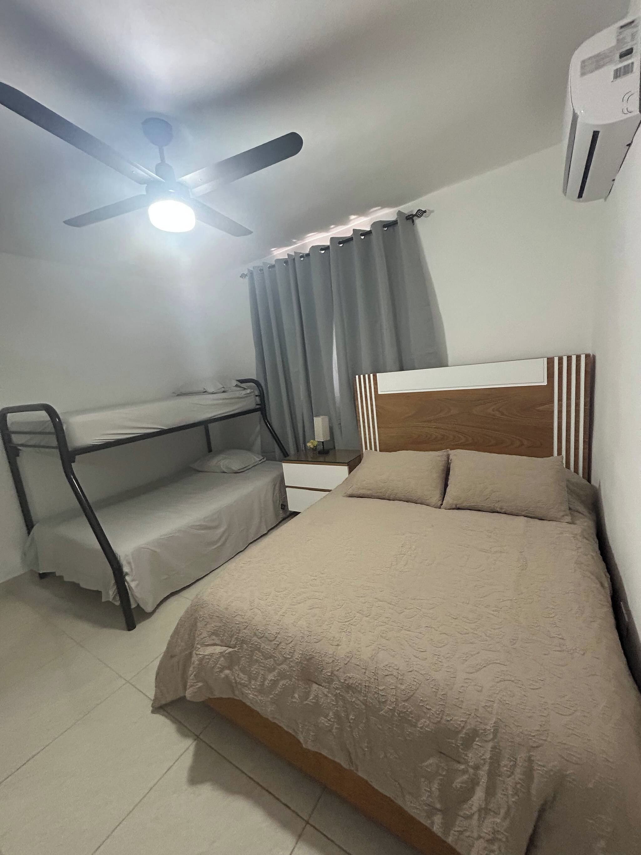 Un dormitorio con una cama individual doble, un escritorio, una cama con cabecera, un ventilador de techo y un equipo de aire acondicionado.