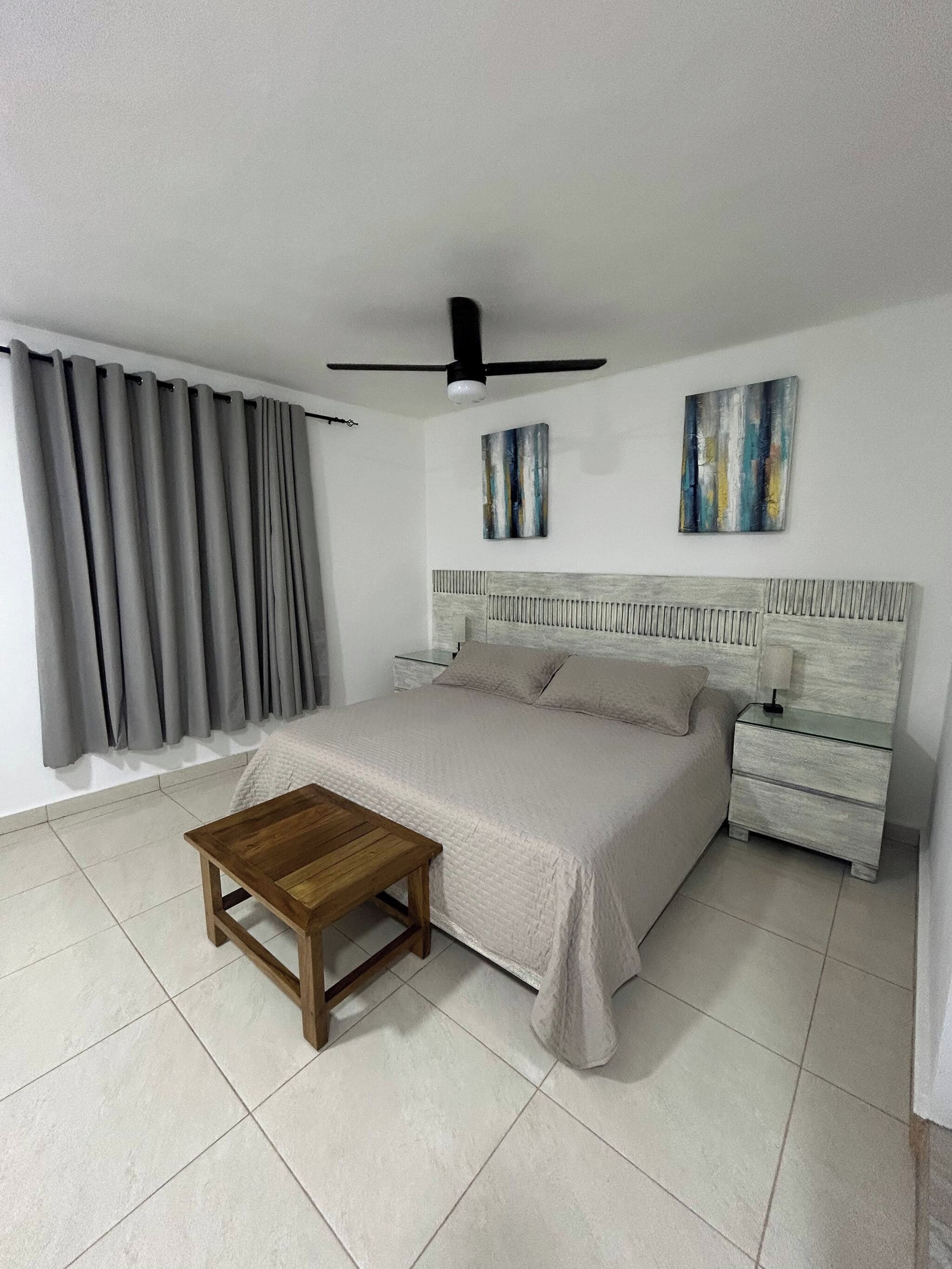 Quarto com cama, mesa de madeira, mesa de cabeceira, ventilador de teto e duas obras de arte nas paredes.
