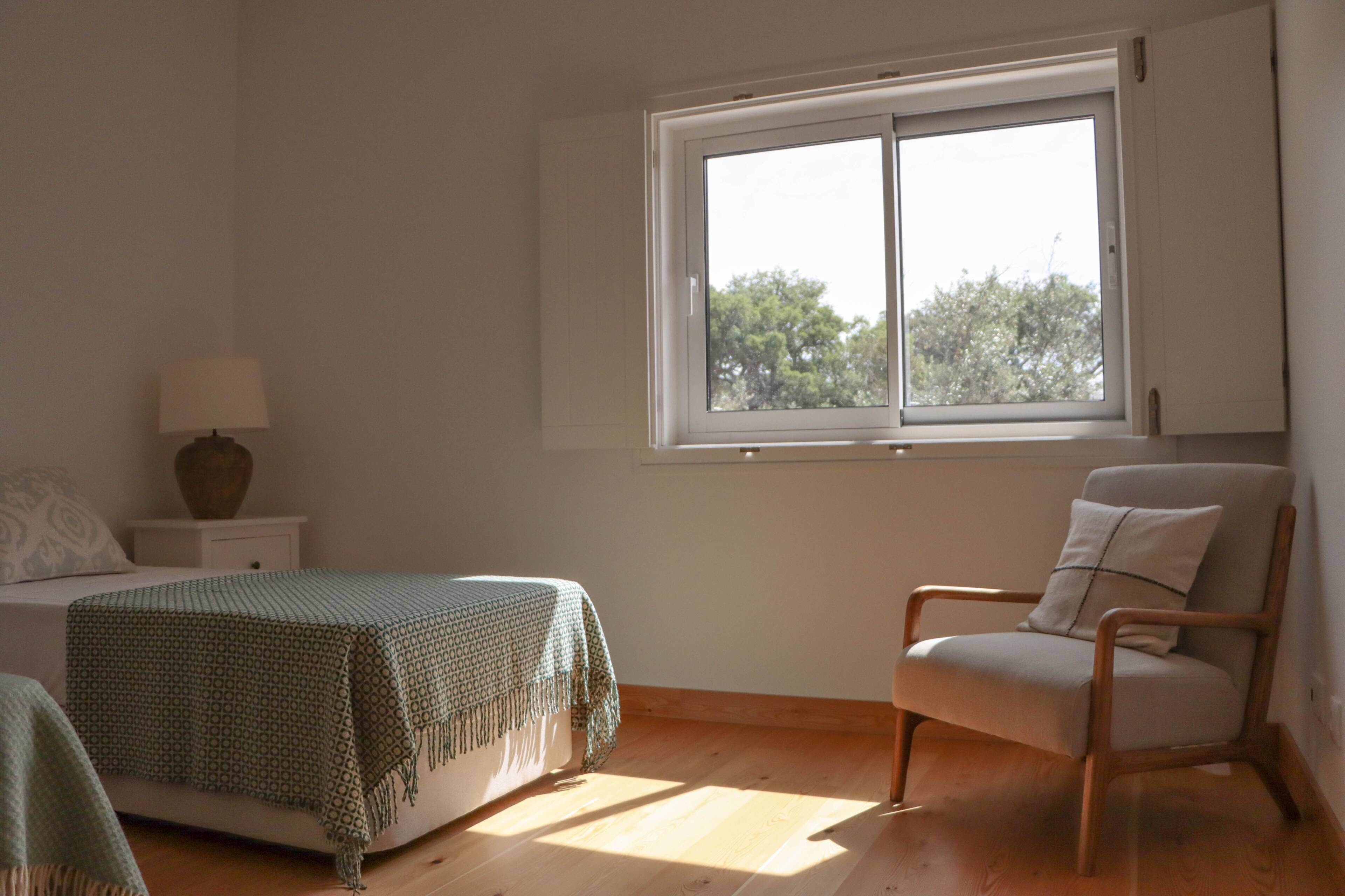 Een slaapkamer met een bed, een stoel, een nachtkastje, een lamp en een raam met uitzicht op bomen.