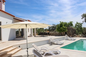 A pool area with lounge chairs and a table, a stone wall, and a building with a tiled roof.