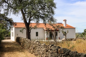 A white building with a red-tiled roof, a stone wall, and a tree in the foreground.