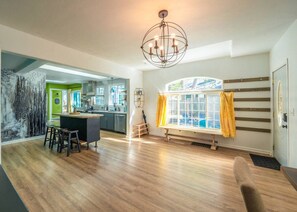 A spacious living area with a wooden floor, a dining area with stools, a large window with curtains, and a kitchen in the background. - Treehouse - Sleeps 14, Hot Tub, 2-Story Solarium (Sioux Falls)