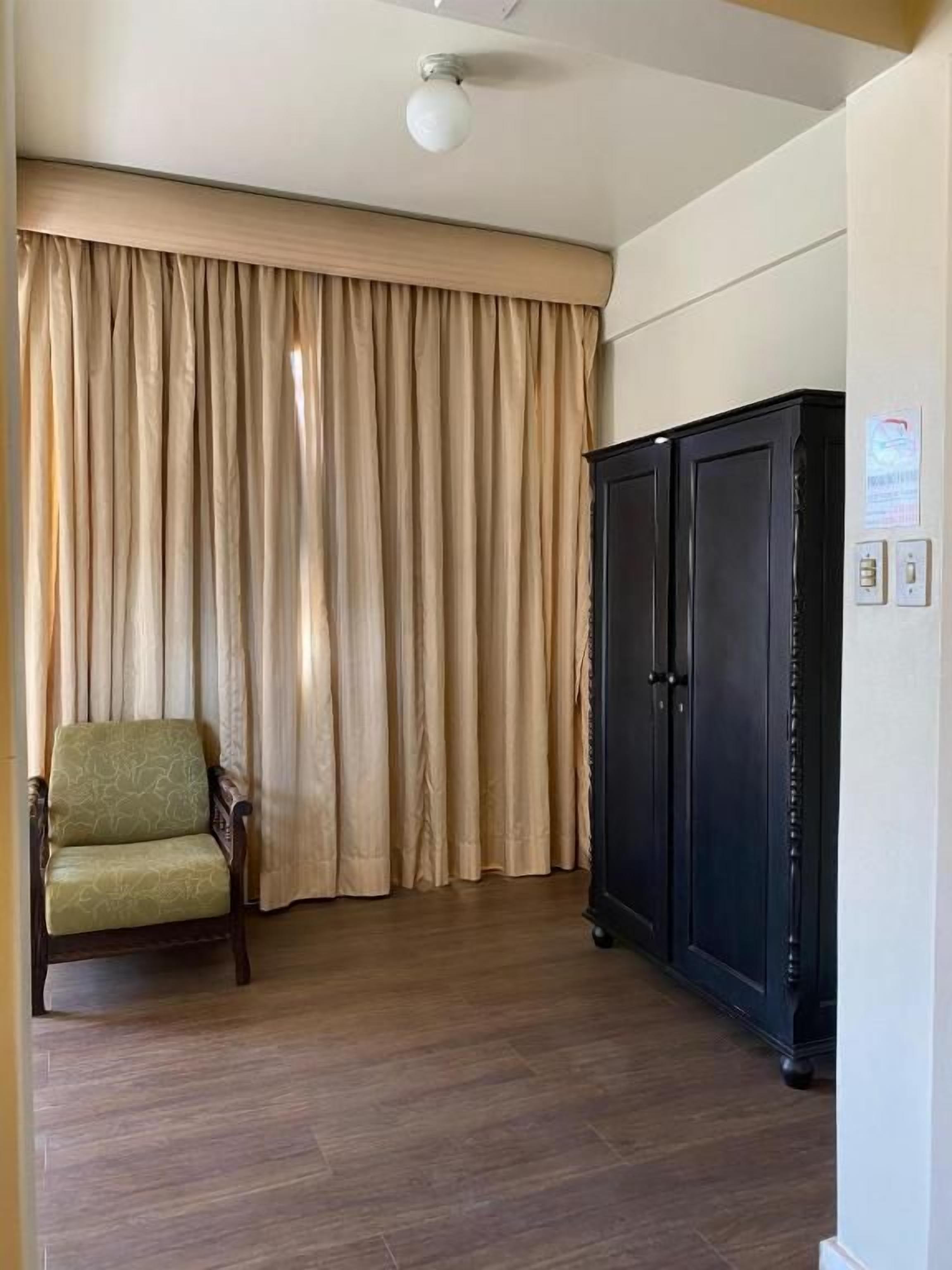 Una habitación con piso de madera, una silla tapizada de verde, un armario negro grande y cortinas color beige.