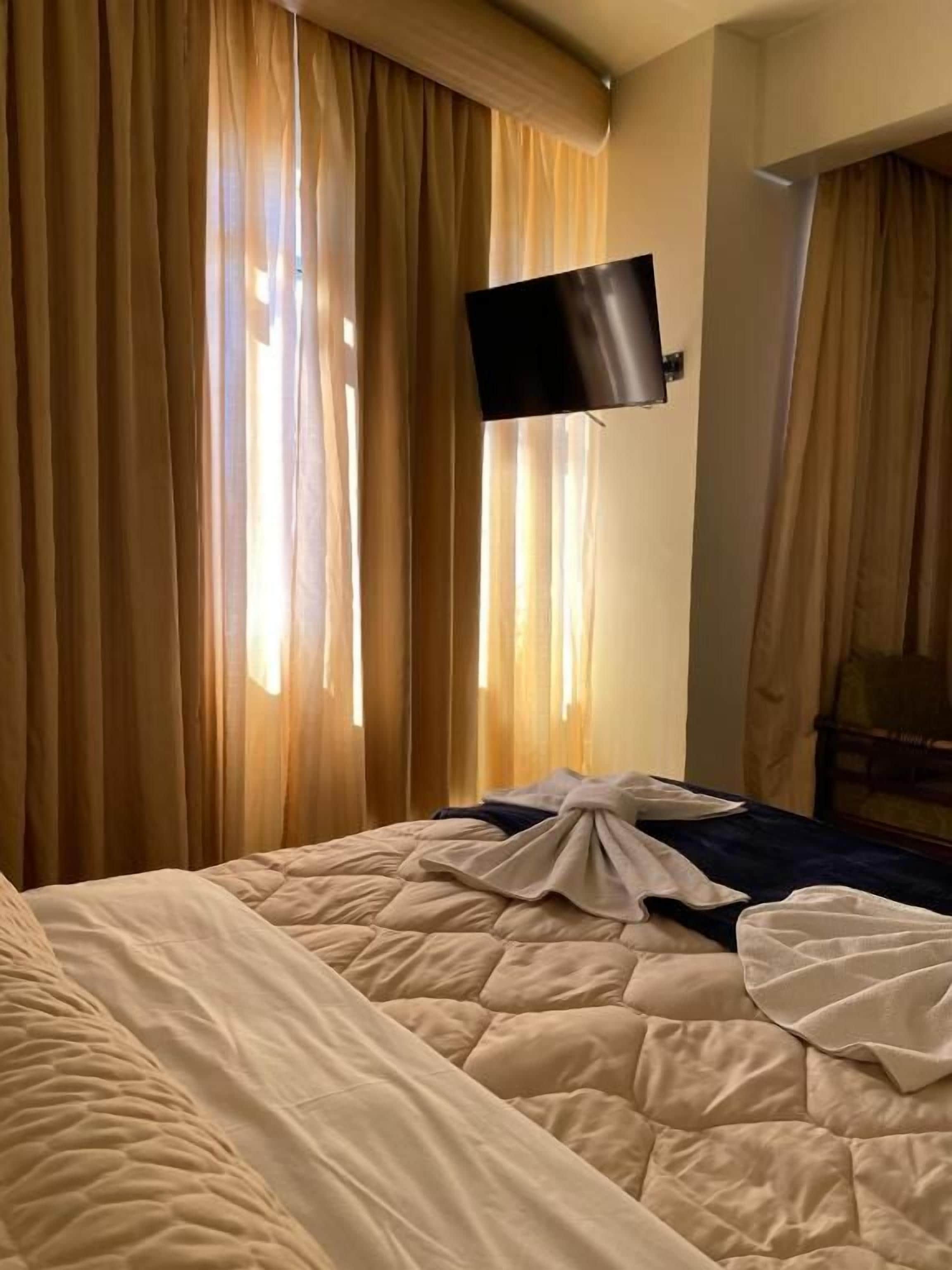Habitación de hotel con cama, cortinas, televisor de pantalla plana montado en la pared y cama bien hecha con manta.