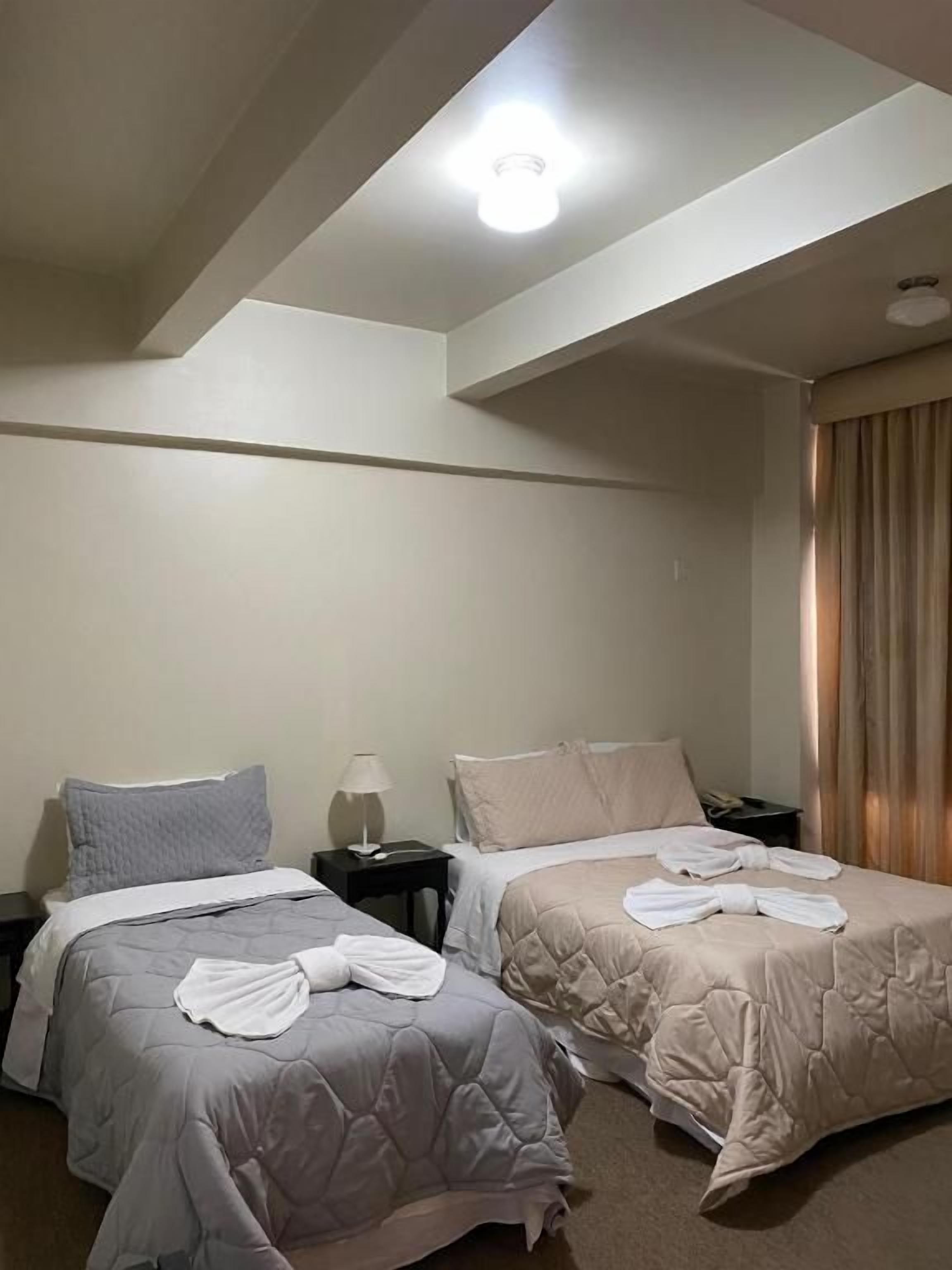 Hotellihuone, jossa on kaksi sänkyä, yöpöytä, lamppu ja kattovalaisin.