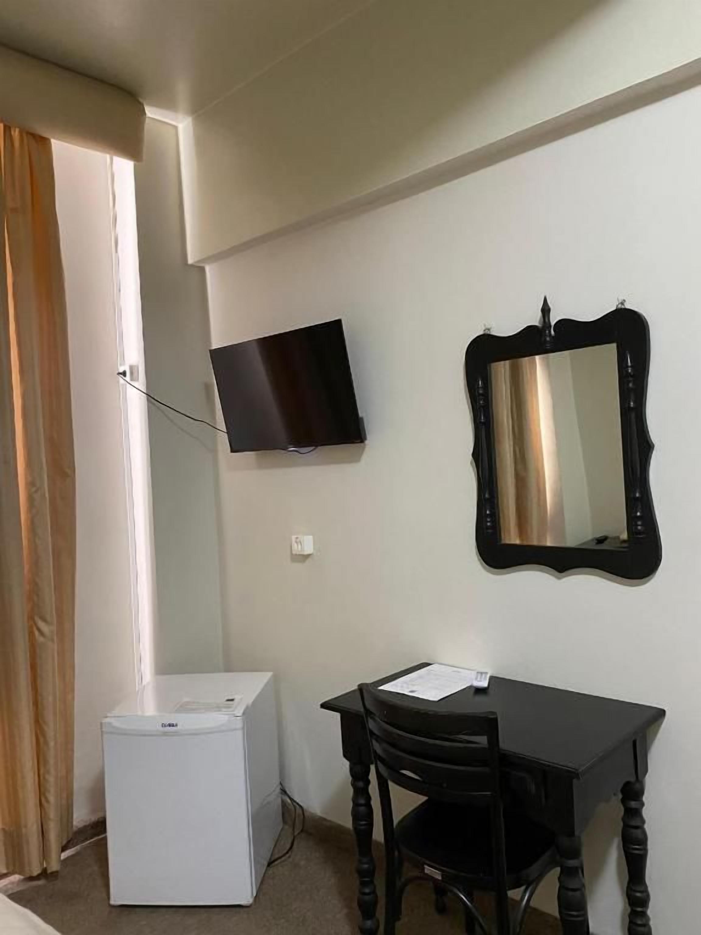 A room with a flat-screen TV mounted on the wall, a small white appliance, a wooden table with a chair, and a mirror with an ornate frame.