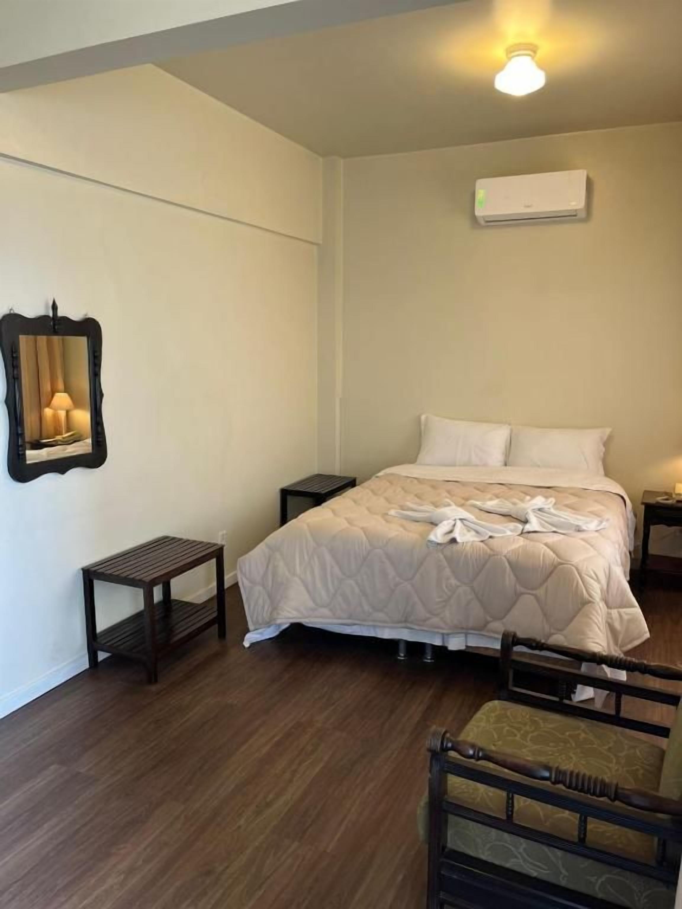 A bedroom with a bed, bedside tables, a nightstand, a chair, a mirror, and an air conditioning unit.
