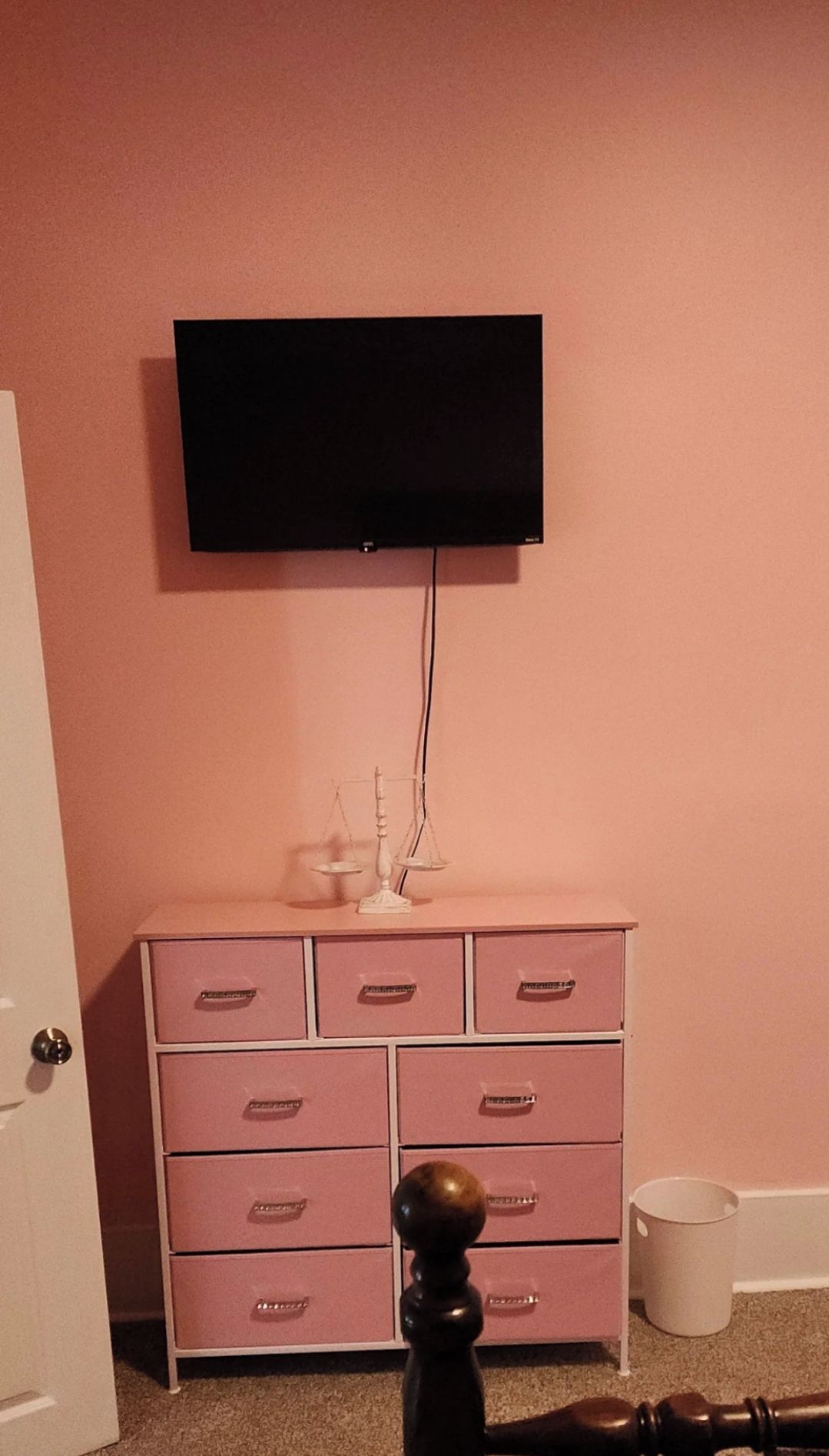 A room with a pink wall, a white dresser with drawers, a flat-screen TV mounted above the dresser, and a door on the left.
