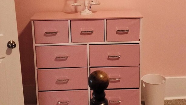 A room with a pink wall, a white dresser with drawers, a flat-screen TV mounted above the dresser, and a door on the left. - 2-bedroom home great for workforce housing, family, and content creation! (Americus)