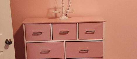 A room with a pink wall, a white dresser with drawers, a television mounted above it, and a wooden bed frame.