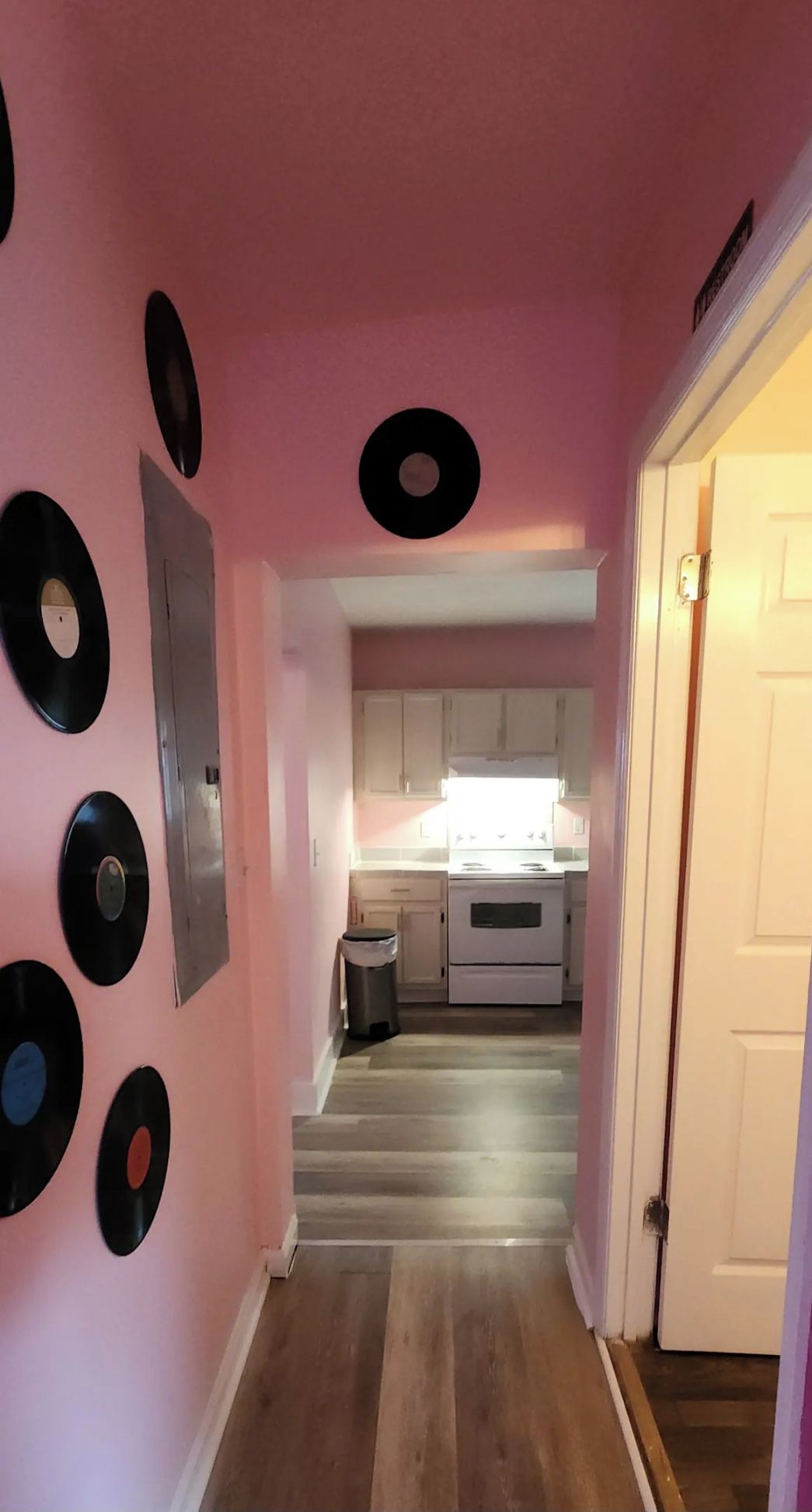 A hallway with pink walls, a wooden floor, and a kitchen area with white cabinets and a stove.