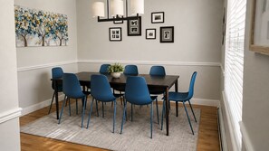 A dining area with a dark wooden table, blue chairs, a grey rug, and a wall with framed photos and a painting. - Spacious 3BR - 2 Kings, Pool Table + Patios (Centennial)