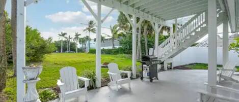 Un espacio al aire libre techado con sillas blancas y una parrilla, con vistas a un jardín y una escalera.