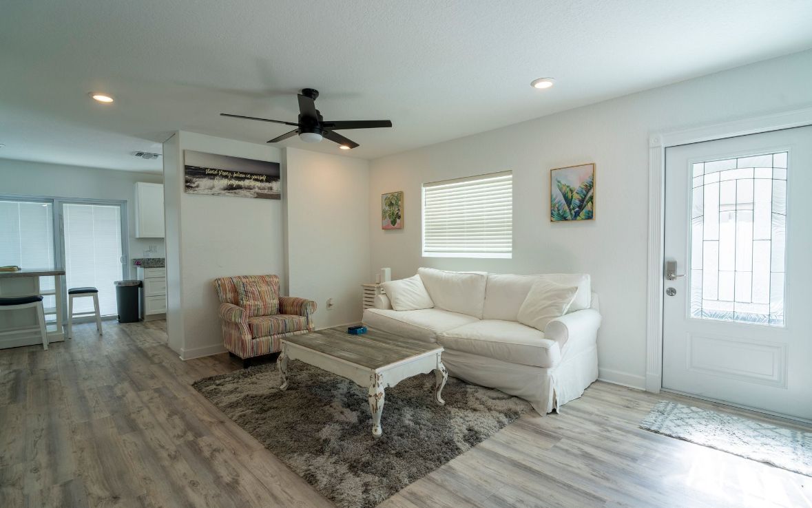 A living room with a white sofa, a patterned armchair, a coffee table, a ceiling fan, and a window with blinds.