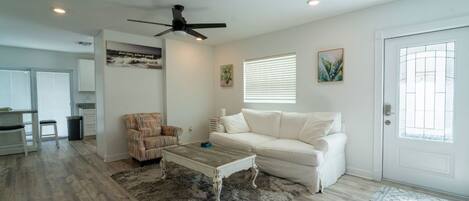 A living room with a white sofa, a patterned armchair, a coffee table, a ceiling fan, and a window with blinds.