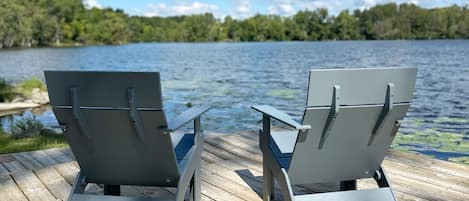 Two Adirondack chairs on a wooden deck overlooking a lake with a forested shoreline.