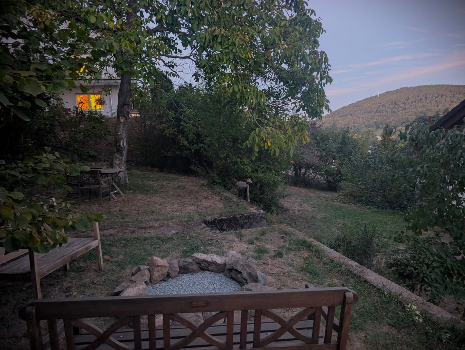 A rustic outdoor seating area with a stone fire pit, wooden benches, and a view of a hillside.