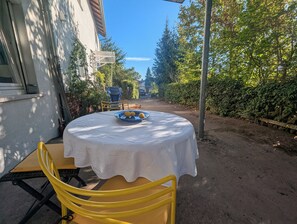 Een ronde tafel met een wit tafelkleed en een blauw bord met citroenen, voor een achtergrond van een onverhard pad en begroeiing.