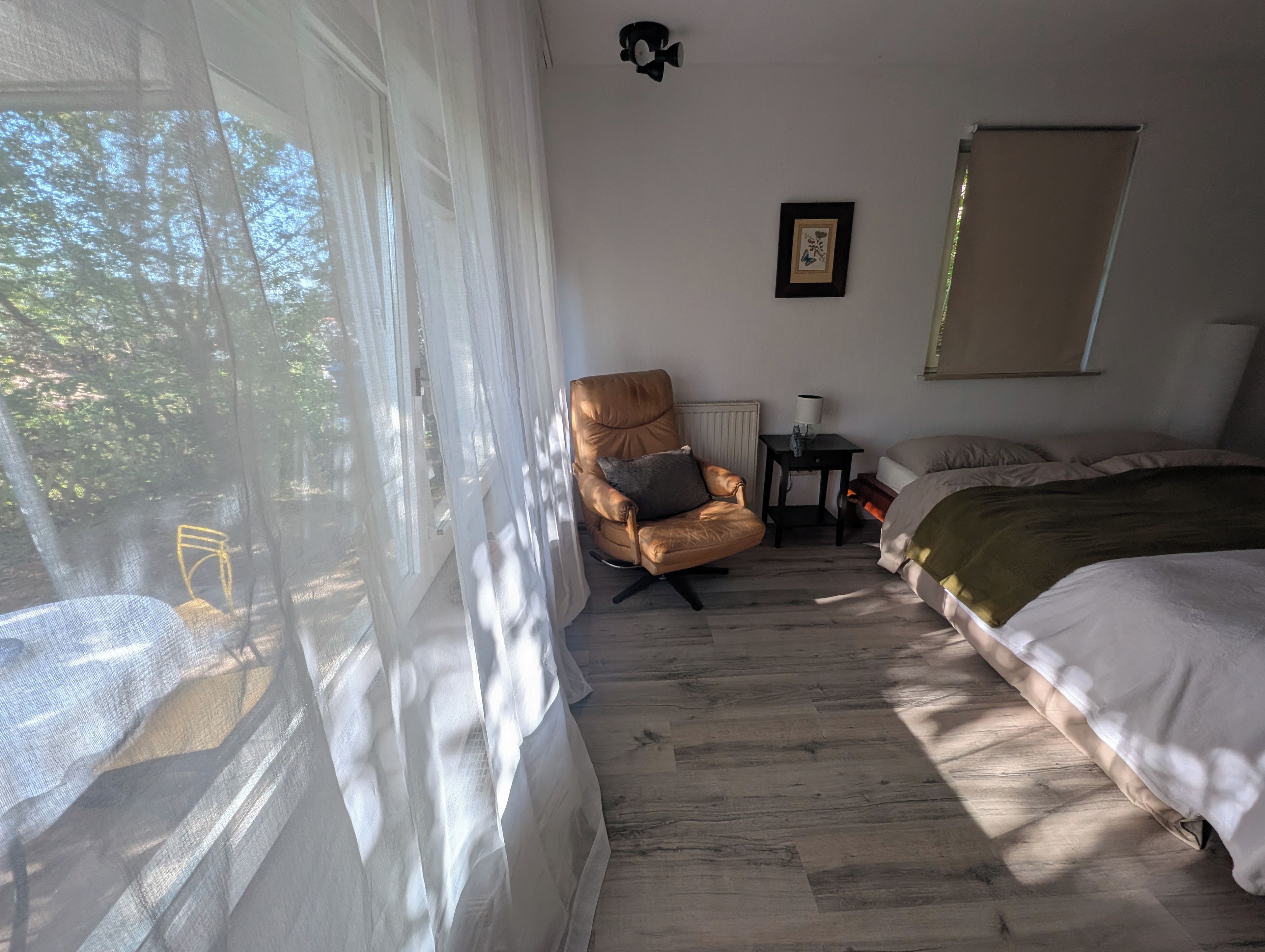 A bedroom with a bed, a chair, a table, and a window with sheer curtains.