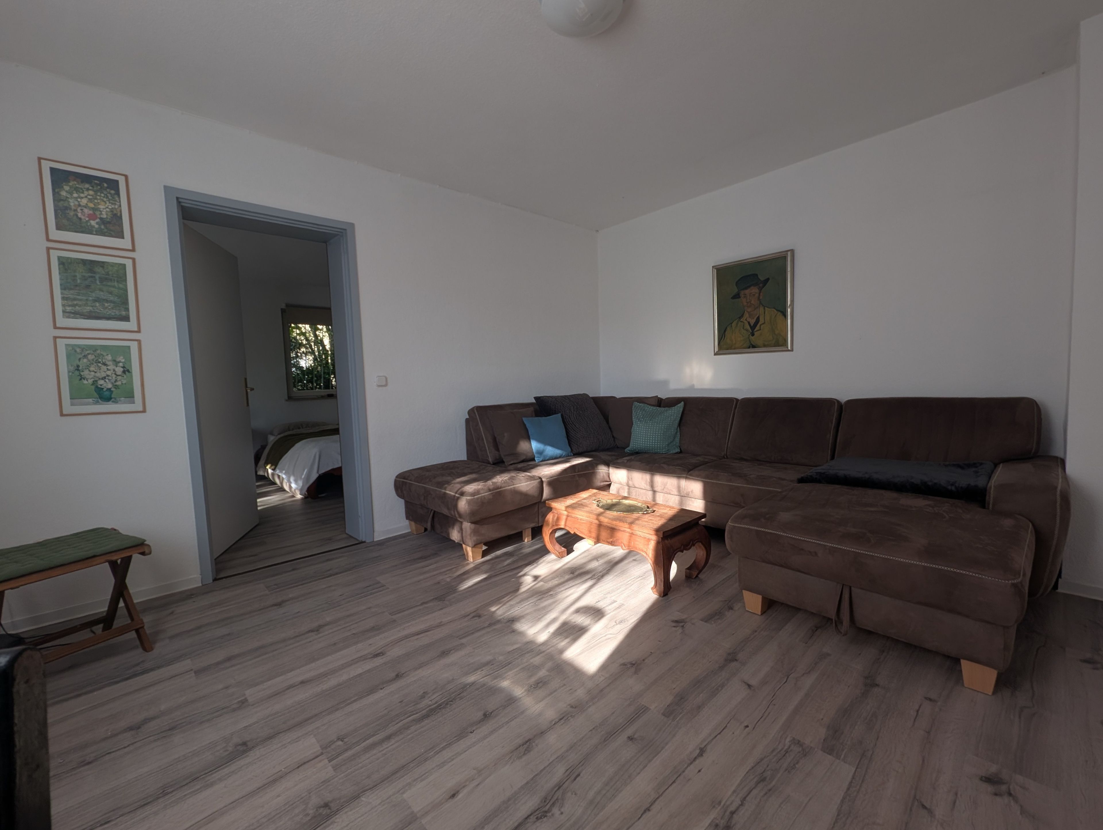 A living room with a brown sofa, a wooden coffee table, and a bench. There are framed pictures on the wall and a painting above the sofa.
