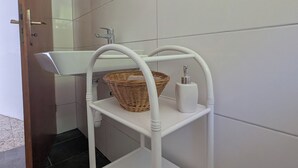 Une salle de bain avec un lavabo blanc, une étagère blanche sur laquelle repose un panier en osier, un distributeur de savon blanc et un miroir surmonté d’une étagère en bois.