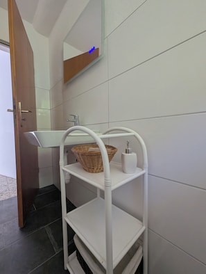 Een badkamer met een witte wastafel, een wit plankje met een rieten mand, een witte zeepdispenser en een spiegel met een houten plank erboven.
