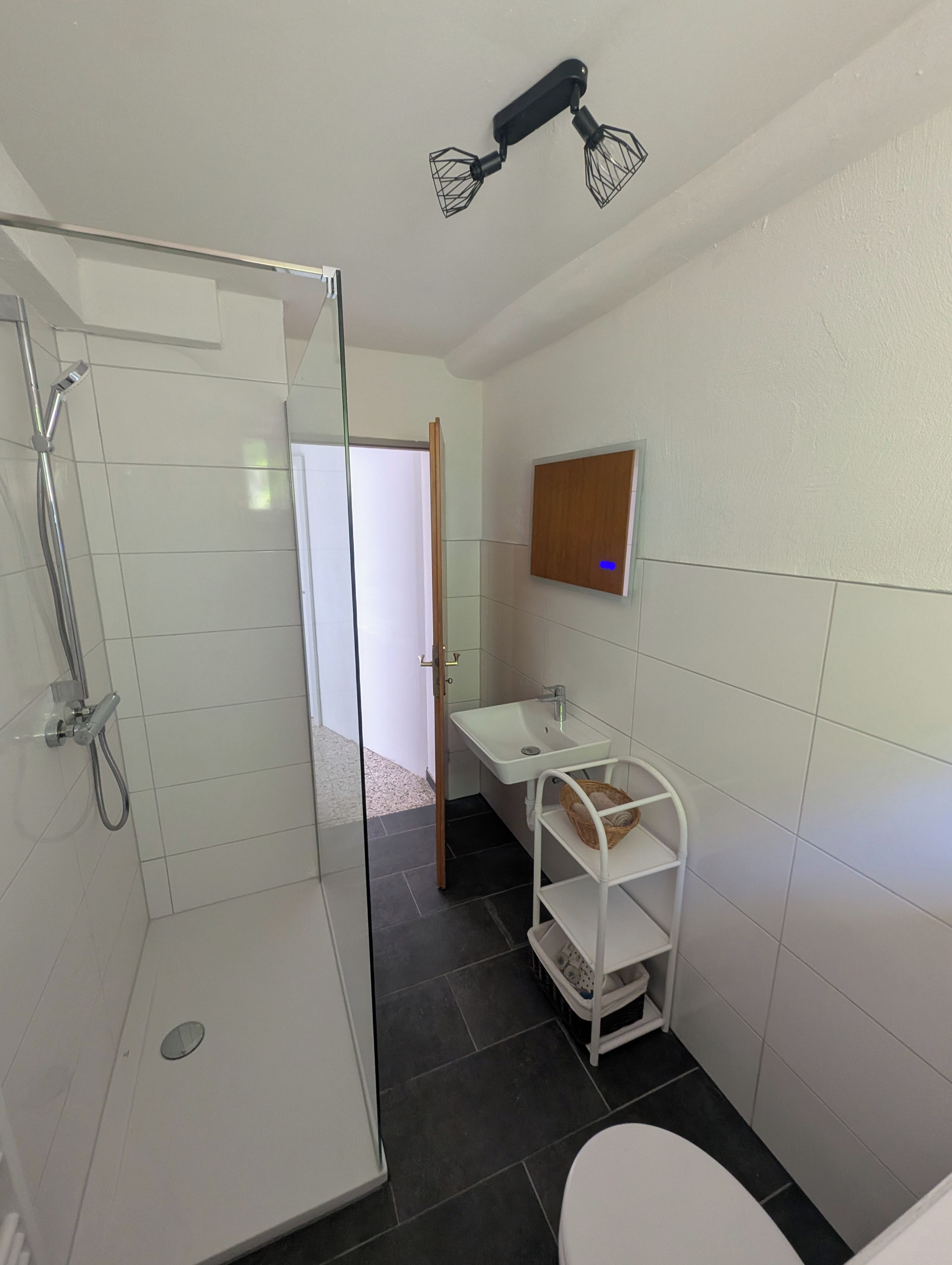 Ein Badezimmer mit einer Duschkabine, einem weißen Waschbecken, einem Spiegel und einem Regal mit einem Korb.