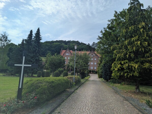 A cobblestone pathway leading to a building with a red-tiled roof, surrounded by lush greenery and a cross-shaped monument. - La Maison sur la Colline – Eco-Friendly Getaway with Garden & Scenic Views (Höchst im Odenwald)