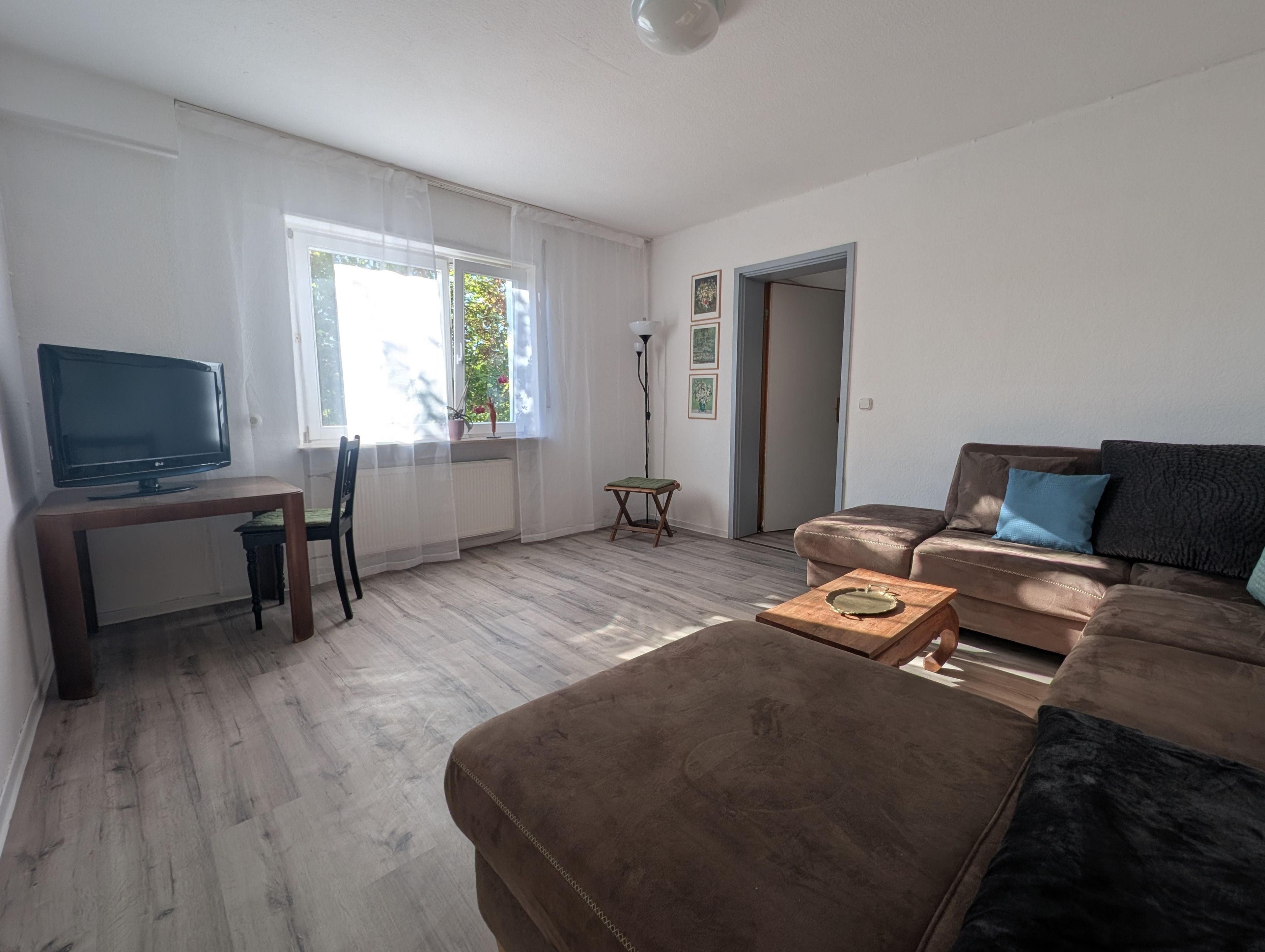 Ein Wohnzimmer mit einer Couch, einem Fernseher, einem kleinen Tisch und einem Sessel. Es gibt ein Fenster mit Vorhängen und eine Heizung.