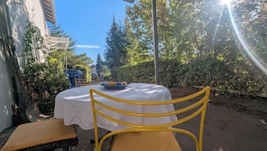 Un espacio al aire libre cubierto con mesa y sillas, rodeado de follaje y una valla.