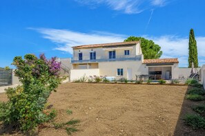 Ein zweistöckiges Haus mit Balkon, Schwimmbad und einem Garten mit violetten Blumen.