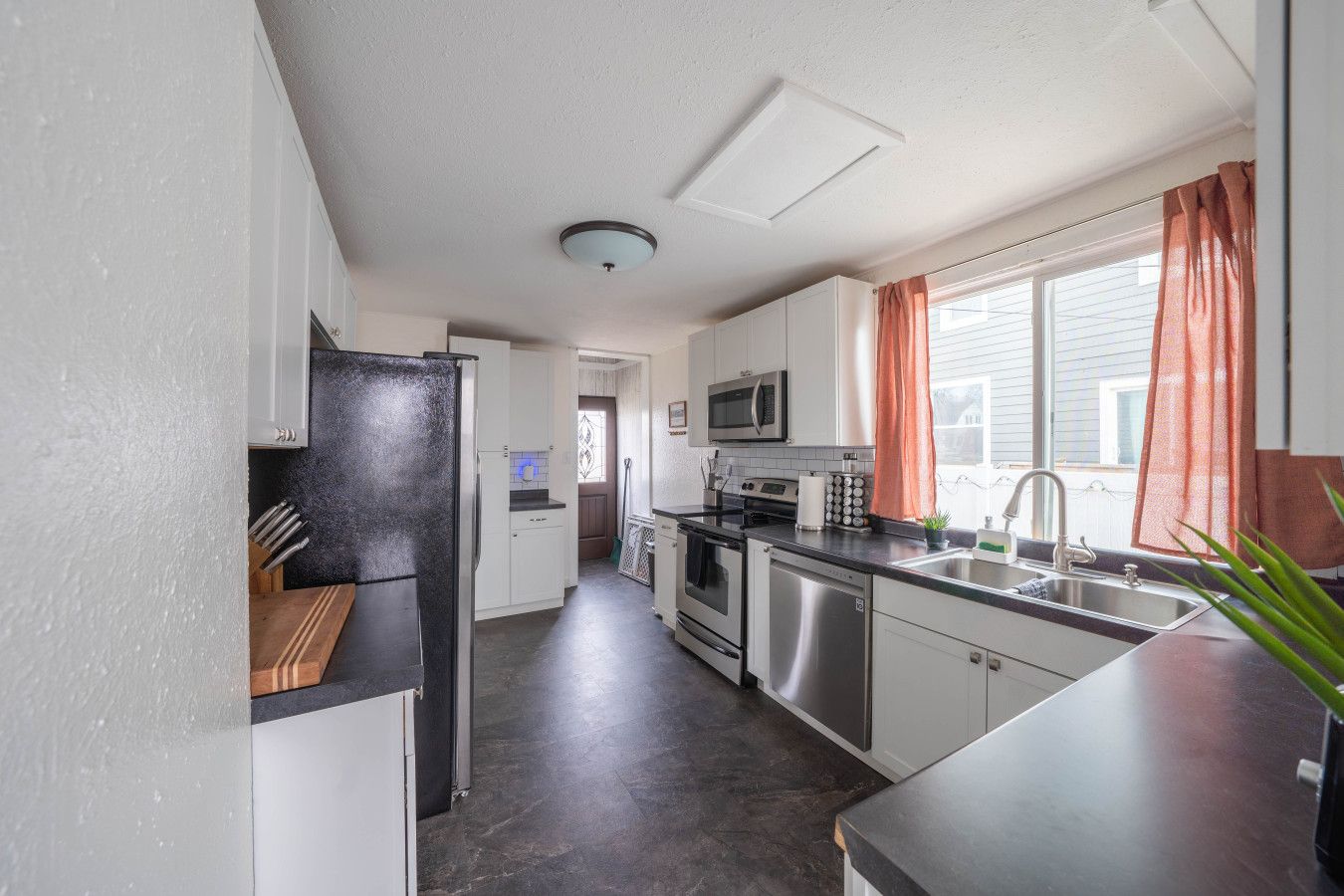 A compact kitchen with white cabinets, stainless steel appliances, and a window with curtains.