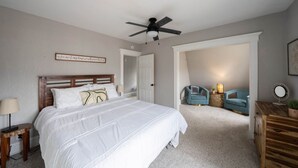 A bedroom with a large bed, a ceiling fan, a chair, a small table, and a mirror. - Mission House: Historic 4BR Near Downtown (Sioux Falls)