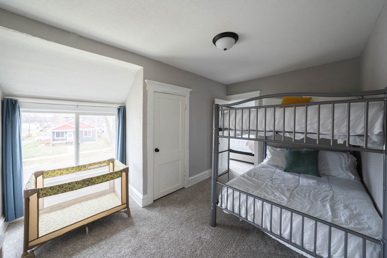A room with a bunk bed, a door, a window with curtains, and a view of houses outside.