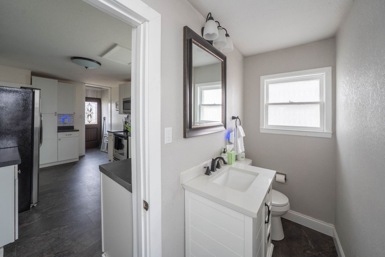 A modern bathroom with a white sink, a toilet, and a mirror above the sink. There is a window on the wall and a door leading to the kitchen.