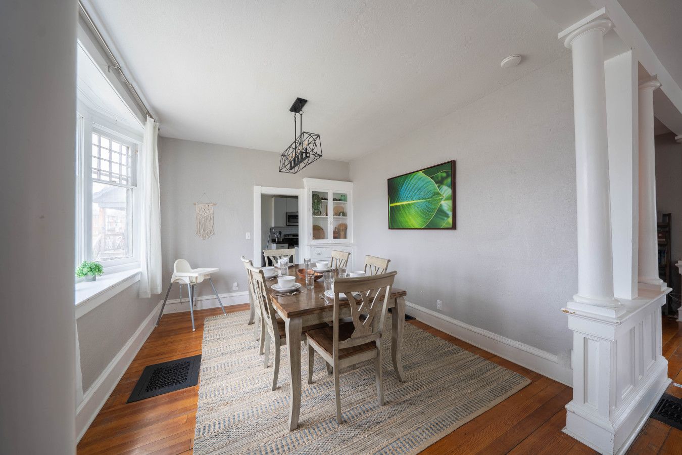 A dining area with a wooden table and chairs, a patterned rug, and a green leaf painting on the wall.