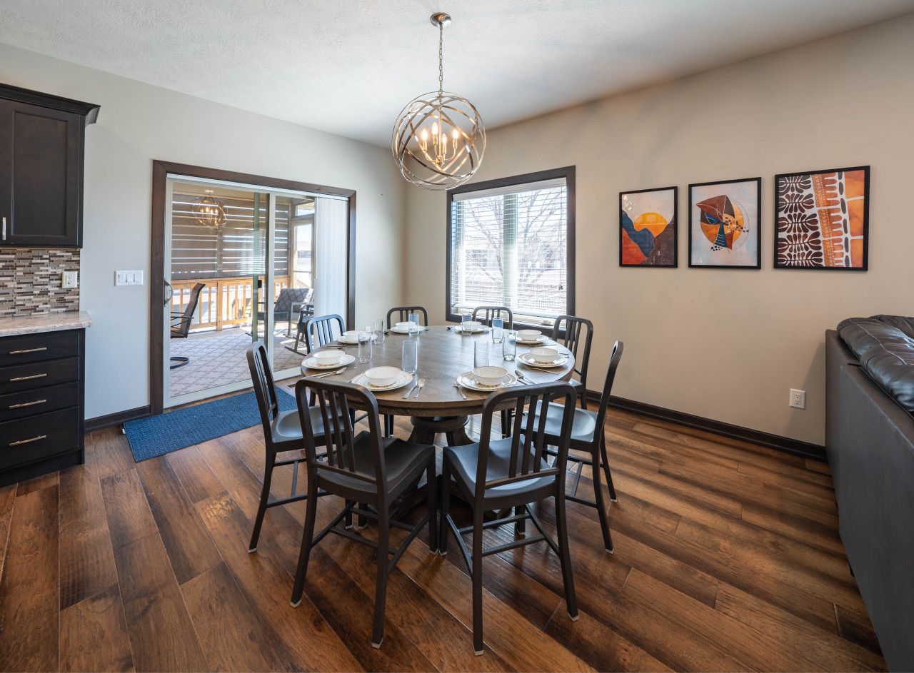 Comedor con mesa redonda dispuesta para seis personas, rodeada de sillas, en una habitación con suelo de madera y un ventanal grande.