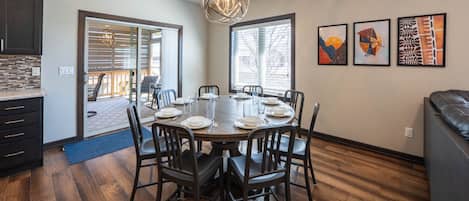 A dining area with a round table set for six, surrounded by chairs, in a room with wooden flooring and a large window.