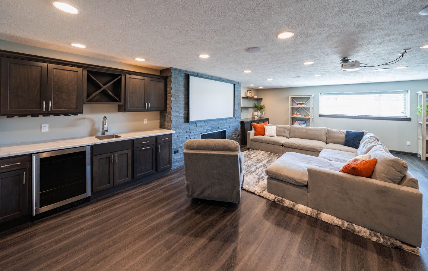 Una cocina moderna con armarios oscuros y una pared de acento de piedra, una zona de estar acogedora con un sofá modular grande y un proyector de techo.