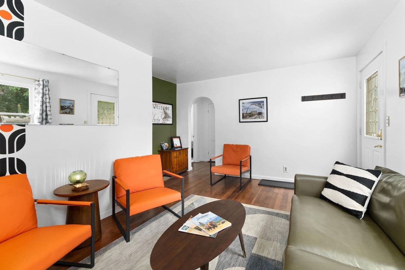 Ein modernes Wohnzimmer mit zwei orangefarbenen Sesseln, einem braunen Couchtisch und einem grünen Sofa mit Kissenmuster.