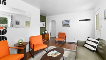 Ein modernes Wohnzimmer mit zwei orangefarbenen Sesseln, einem braunen Couchtisch und einem grünen Sofa mit Kissenmuster.