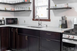 A modern kitchen with dark cabinetry, a white countertop, a stainless steel sink, a microwave, a gas stove, and overhead shelves with kitchenware.