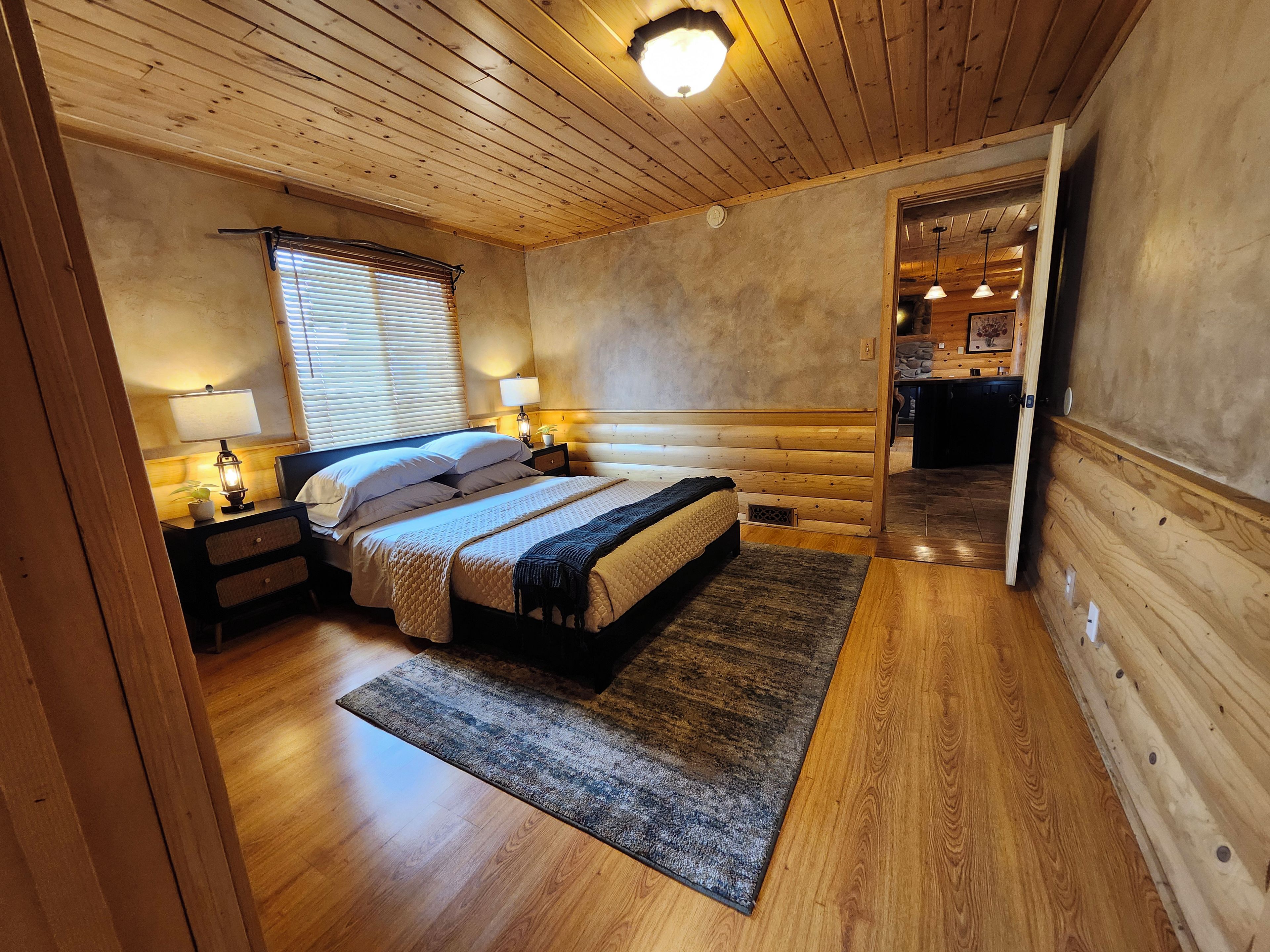 A wooden-paneled bedroom with a bed, bedside lamps, and a view into the kitchen.