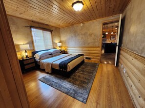 A wooden-paneled bedroom with a bed, bedside lamps, and a view into the kitchen.