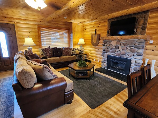 A log cabin interior with a stone fireplace, wooden furniture, and a television mounted on the wall.