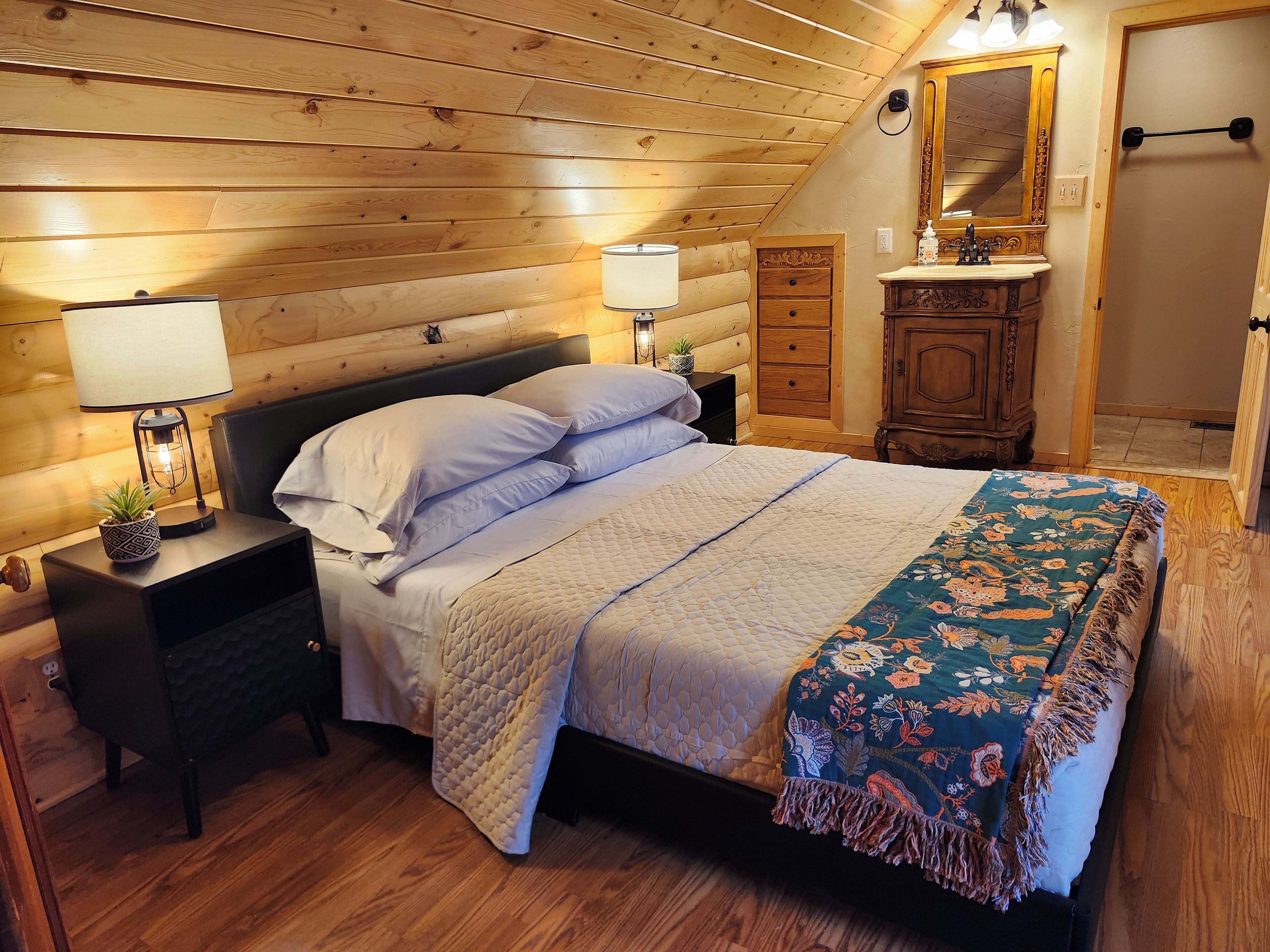 A bedroom with a wooden ceiling, a bed with white bedding, a nightstand with a lamp, a dresser, and a mirror.