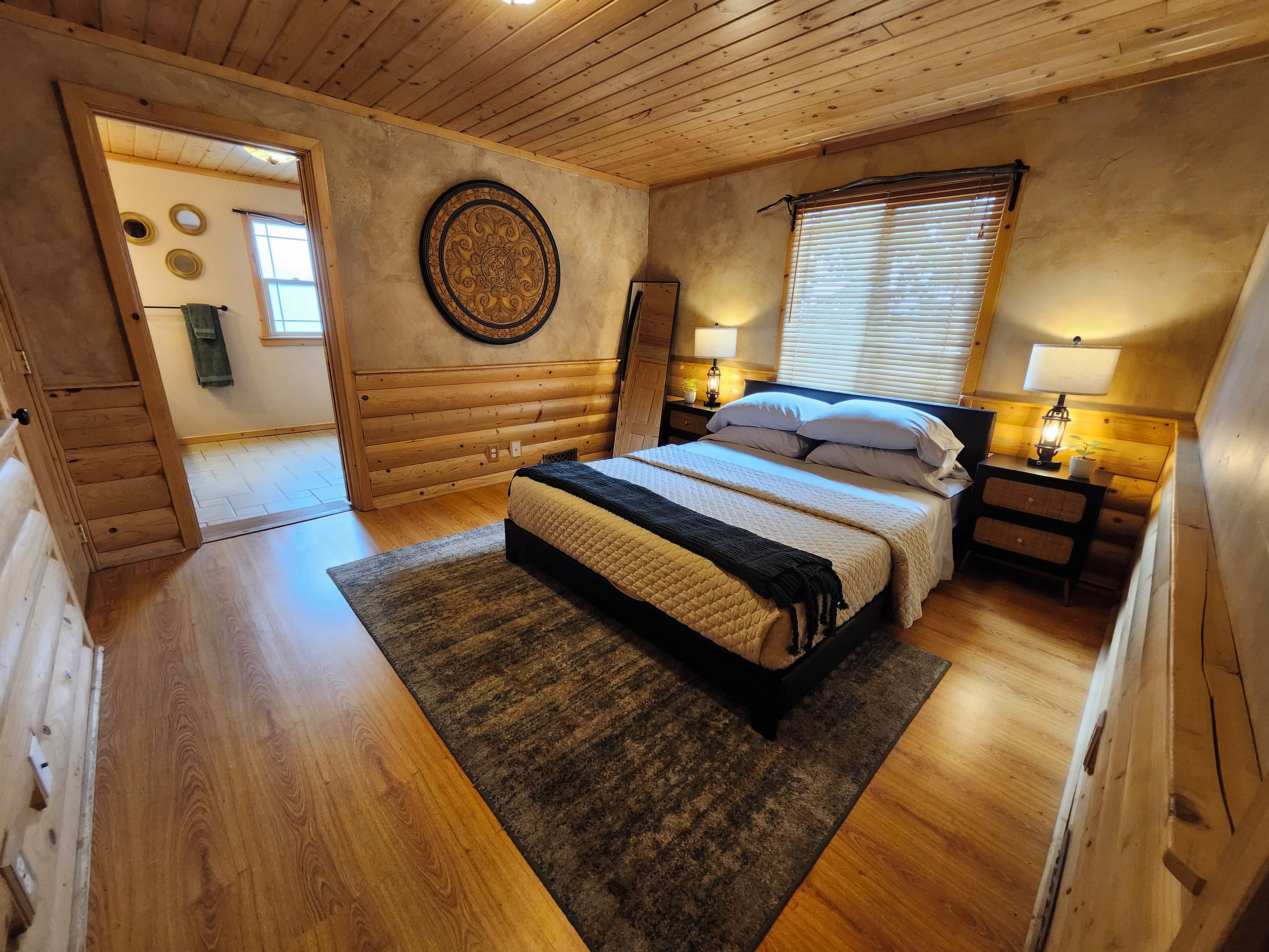 A bedroom with a wooden bed, a nightstand with a lamp, a circular wall decoration, and a window with blinds.