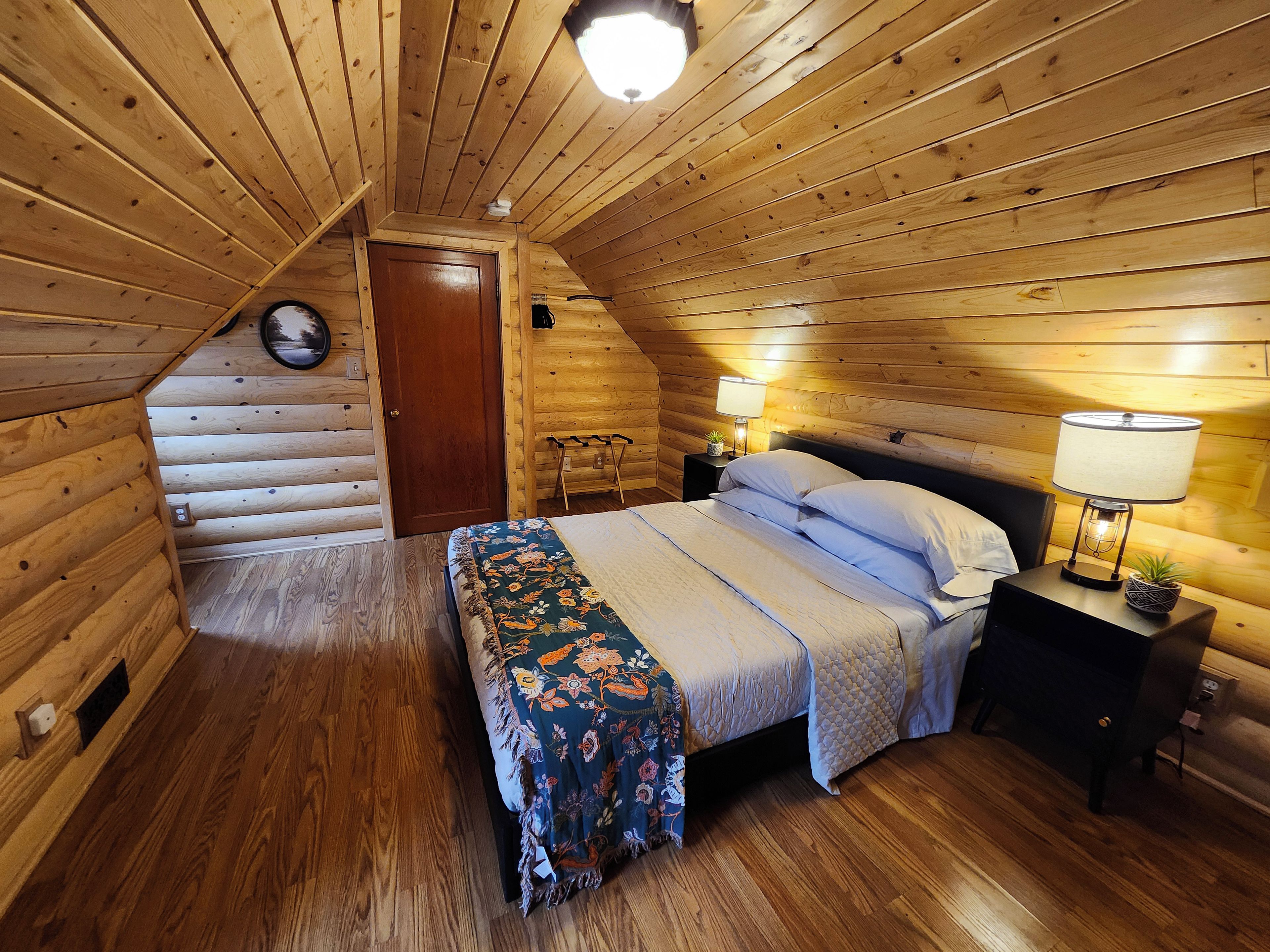 Un dormitorio acogedor tipo cabaña de madera, con cama, lámparas de noche y techo de madera.
