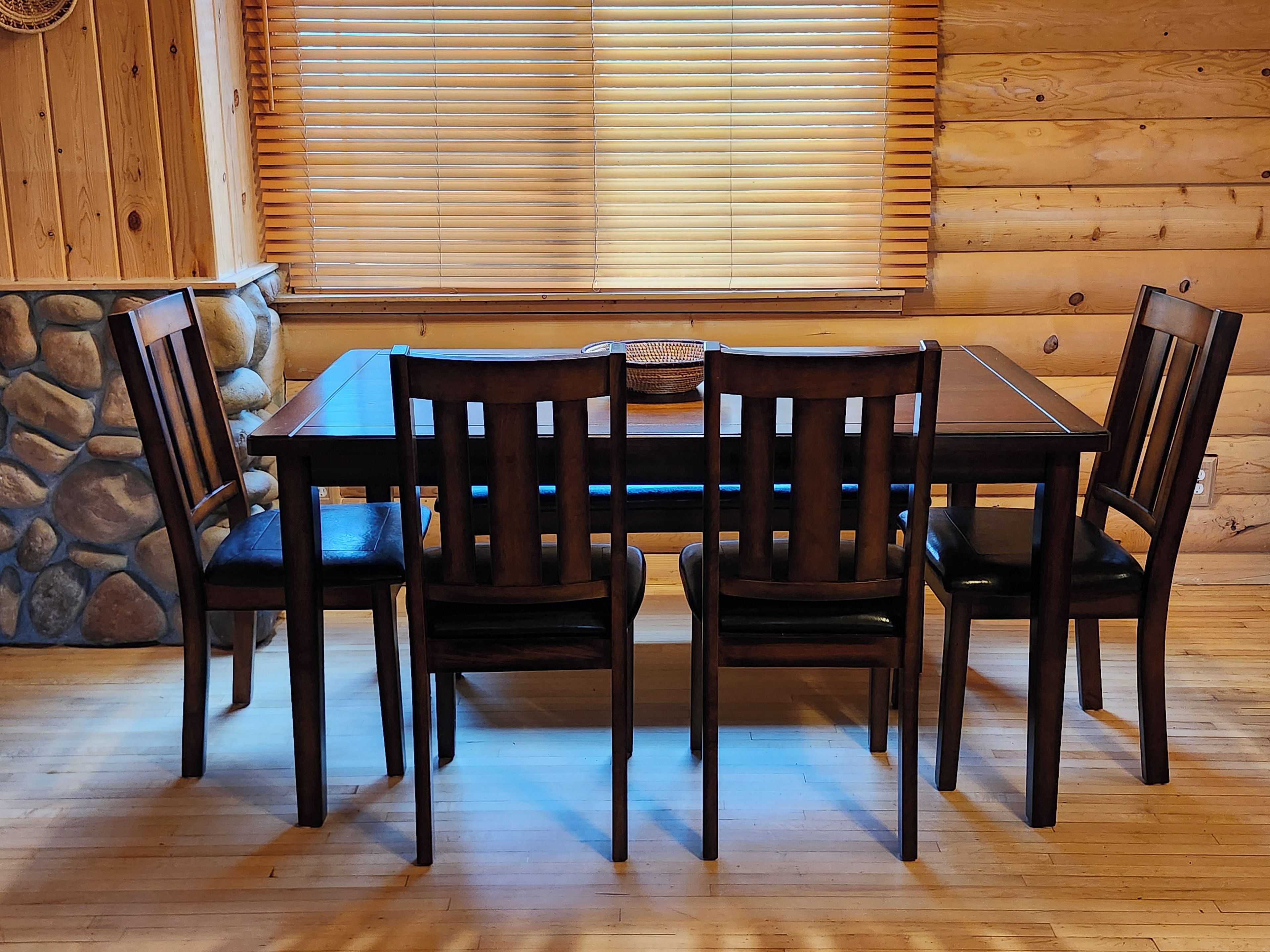 A wooden dining table with four chairs, set against a stone wall and wooden blinds.